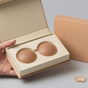 May include: Two round, beige silicone nipple covers housed in a cream-colored molded insert within an open, light tan rectangular box. A matching closed tan box with a pull tab sits beside the open box, accompanied by four small, light-colored petals.