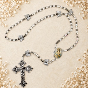 May include: A rosary featuring clear and metallic gray beads interspersed with silver-toned butterfly charms. The piece includes an oval centerpiece depicting a religious figure and a detailed, ornate silver-toned crucifix at the bottom end.