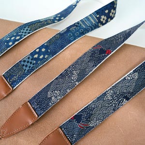 May include: Four patterned camera straps with brown leather ends. The straps feature various blue and white designs, including floral and wave patterns. The straps have a white backing and are arranged diagonally across the image.