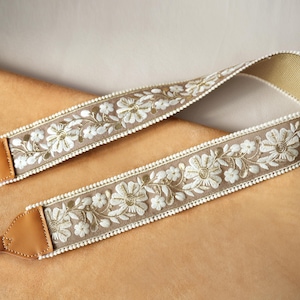 May include: A beige camera strap with white floral embroidery and gold accents. The strap features brown leather ends and a delicate white beaded trim along the edges. The design is symmetrical, with the floral pattern repeating along the length of the strap.