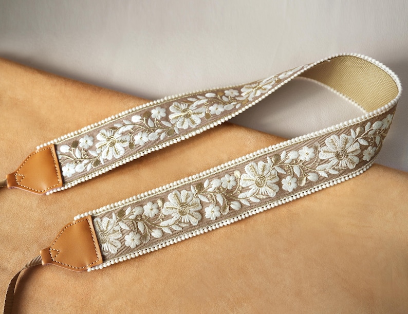 May include: A beige camera strap with white floral embroidery and gold accents. The strap features brown leather ends and a delicate white beaded trim along the edges. The design is symmetrical, with the floral pattern repeating along the length of the strap.
