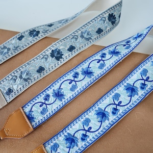 May include: Three camera straps with floral embroidery. The straps feature a white background with blue floral patterns and accents. The ends are made of tan leather. The straps are designed for cameras.