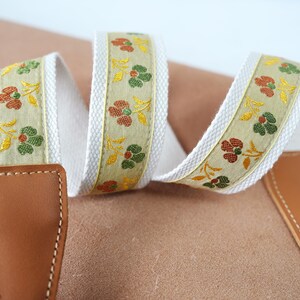 Puede incluir: Una correa de cámara con una base de color crema y un estampado floral en tonos verdes, naranjas y amarillos. La correa tiene extremos de cuero marrón con costuras blancas.