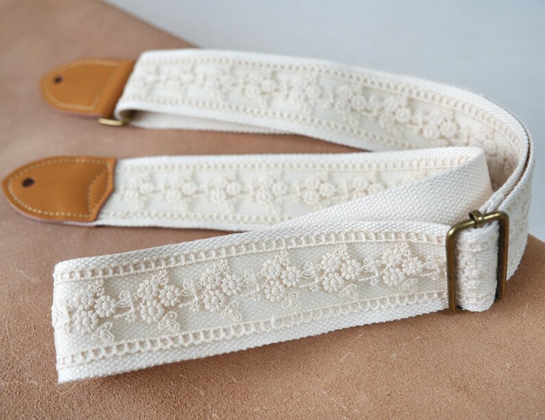 May include: Cream guitar strap with floral lace trim. It has tan leather ends and a brass buckle. The design blends vintage style with elegance, suitable for acoustic or electric guitars. The strap measures approximately 150 cm in length.