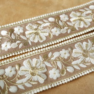 May include: Two strips of decorative ribbon with a tan background. The ribbon features white embroidered floral designs with gold accents. The edges are trimmed with small white pom-poms, creating a delicate and elegant look. Ideal for craft projects.