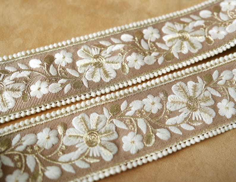 May include: Two strips of decorative ribbon with a tan background. The ribbon features white embroidered floral designs with gold accents. The edges are trimmed with small white pom-poms, creating a delicate and elegant look. Ideal for craft projects.