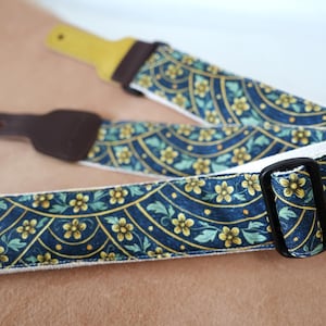 Op de afbeelding: Een gitaarband met een donkerblauwe achtergrond en een patroon van gele bloemen en groene bladeren. De band heeft een witte rand en een zwarte verstelbare gesp.