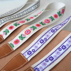 May include: Close-up of several camera straps with floral and geometric patterns. The straps are white with colorful embroidered designs in pink, green, and purple. Each strap has a brown leather end piece.