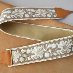 May include: A camera strap with a floral embroidered design. The strap features a tan background with white and gold flowers and green leaves. The edges are trimmed with a white, scalloped trim. The ends are tan leather.