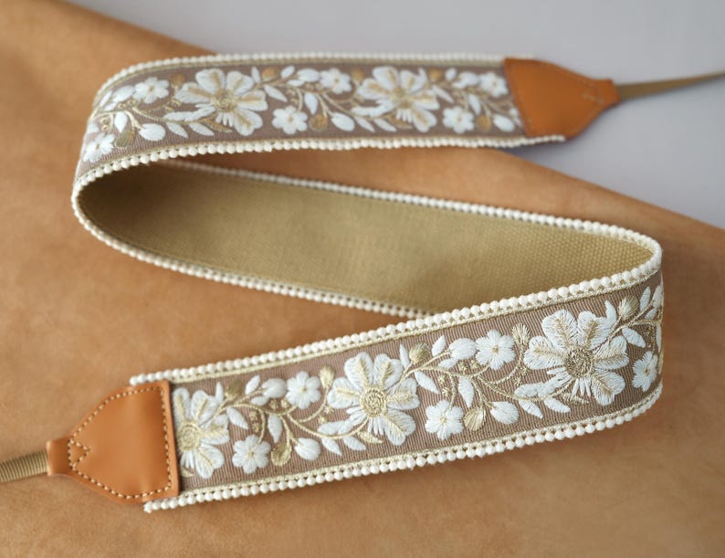 May include: A camera strap with a floral embroidered design. The strap features a tan background with white and gold flowers and green leaves. The edges are trimmed with a white, scalloped trim. The ends are tan leather.