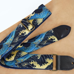 Peut inclure: Une sangle de guitare avec un motif de vagues bleu et or. La sangle a des extrémités en cuir noir et une boucle en métal doré. Le motif de vagues rappelle l'art japonais.