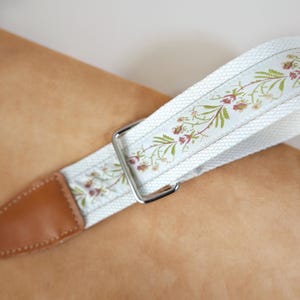 May include: A camera strap featuring a white woven fabric band with a delicate floral pattern in shades of green, pink, and brown. The strap includes a silver metal rectangular adjustment buckle and a tan leather end piece with visible stitching.