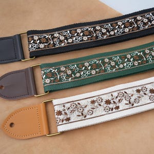 May include: Three guitar straps with floral embroidery. The straps are black, green, and white, with brown and white floral patterns. Each strap has a leather end and gold-coloured metal hardware.