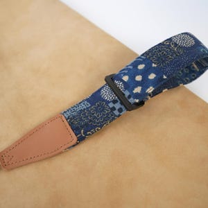 May include: A camera strap featuring a blue fabric section with a white and gold floral and geometric pattern, a black plastic adjustable buckle, and a tan leather end tab with a thin beige nylon webbing strap attached to the end.