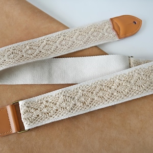 Op de afbeelding: Crèmekleurige gitaarband met een bloemen kanten ontwerp. De band heeft een canvas achterkant en leren uiteinden met een gat voor bevestiging. De metalen hardware is messingkleurig.