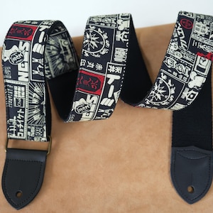 May include: A black guitar strap featuring a repeating pattern of Japanese text and symbols in white and red. The strap has black leather ends and a metal buckle. The strap is approximately 5 cm wide.