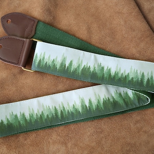 May include: A guitar strap featuring a forest scene design. The strap has a white background with a green forest print, green canvas backing, and brown leather ends. The strap is approximately 2 inches wide.