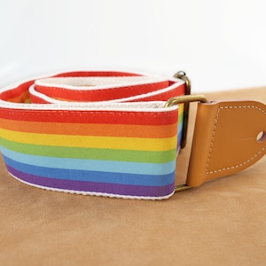 Könnte beinhalten: Ein Gitarrengurt mit Regenbogenmuster. Der Gurt besteht aus Stoff mit einem Farbspektrum, darunter Rot, Orange, Gelb, Grün, Blau und Lila. Er hat ein hellbraunes Lederende und goldfarbene Metallbeschläge.