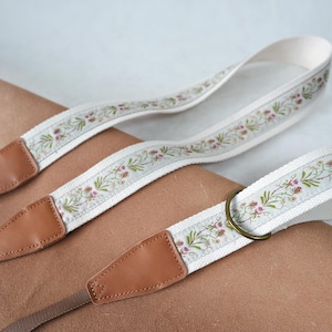 Könnte beinhalten: Zwei Kameragurte mit floralem Design. Die Gurte sind weiß mit einem sich wiederholenden Muster aus grünen Blättern und rosa Blüten. Die Enden sind aus braunem Leder mit einem genähten Design. Ein Gurt hat einen Messingring.