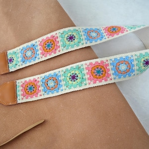 May include: A colorful camera strap with a crocheted design. The strap features a repeating pattern of square motifs in pastel shades of pink, blue, green, and orange, with a cream-colored background. The ends are made of brown leather.