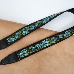 Könnte beinhalten: Schwarzer Kameragurt mit floralem Design. Der Gurt hat ein sich wiederholendes Muster aus blauen Blumen und grünen Blättern, die auf einem schwarzen Hintergrund gestickt sind. Die Enden sind aus schwarzem Leder mit einem schwarzen Riemen zur Befestigung.
