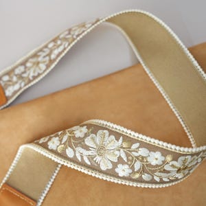 May include: A tan camera strap with a floral embroidered design in white and gold. The strap has a brown leather end piece and white trim along the edges. The strap is made of a woven material.