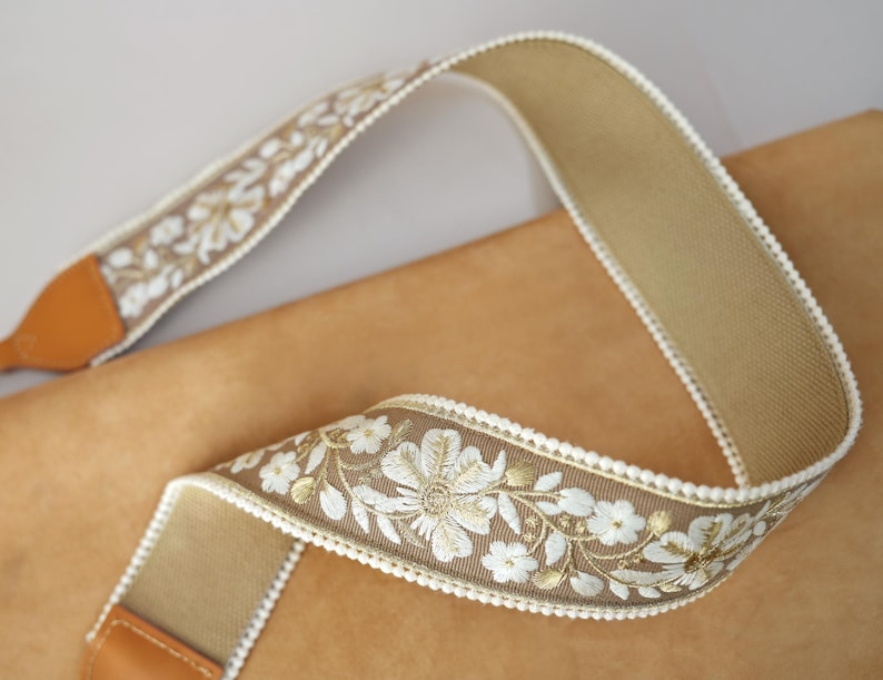 May include: A tan camera strap with a floral embroidered design in white and gold. The strap has a brown leather end piece and white trim along the edges. The strap is made of a woven material.
