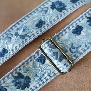 以下が含まれることがあります： 淡い青と白の花柄刺繍ストラップのクローズアップ。ストラップは濃い青色の花と市松模様が特徴です。長方形のアンティーク真鍮色のバックルがストラップに取り付けられています。