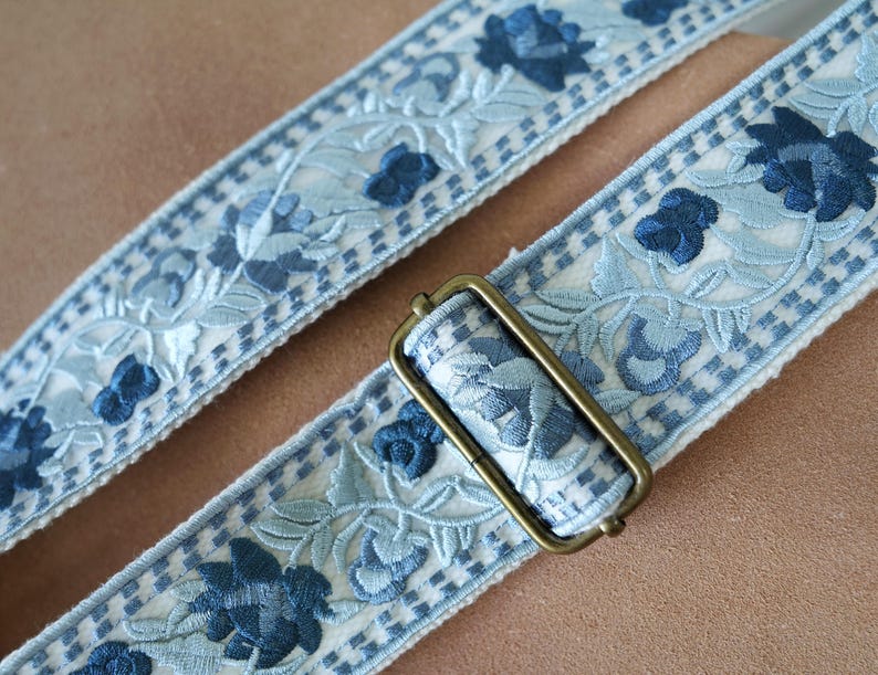以下が含まれることがあります： 淡い青と白の花柄刺繍ストラップのクローズアップ。ストラップは濃い青色の花と市松模様が特徴です。長方形のアンティーク真鍮色のバックルがストラップに取り付けられています。