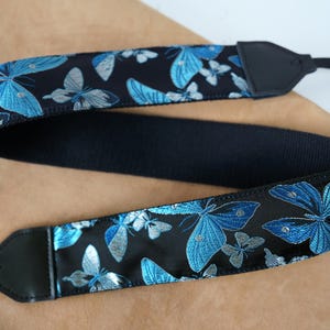 May include: A black camera strap with a repeating pattern of blue and silver butterflies. The strap has black leather ends and a black woven center section. The butterflies have detailed wing patterns.