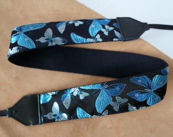 May include: A black camera strap with a repeating pattern of blue and silver butterflies. The strap has black leather ends and a black woven center section. The butterflies have detailed wing patterns.