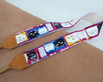 May include: A white camera strap with a colorful cat print design. The strap features black, white, pink, blue, and yellow cats with different expressions. The strap has brown leather ends.