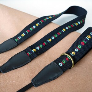 May include: Three black camera straps with colorful embroidered details. The straps have a repeating pattern of small, multi-colored shapes. Each strap has black leather ends and a metal ring on one. The straps are on a light brown surface.