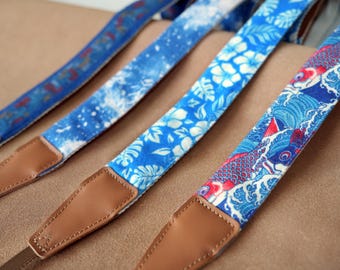 May include: Four blue camera straps with various patterns, including floral, abstract, and koi fish designs. Each strap has brown leather ends and a brown nylon strap. The straps are in a row, angled towards the viewer.
