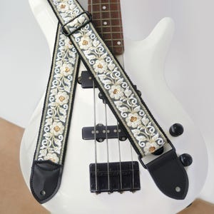May include: A guitar strap featuring a dark gray fabric base with an intricate white and gold floral embroidery pattern. The strap includes black leather ends with circular holes and a black plastic adjustable slider buckle for length adjustment.