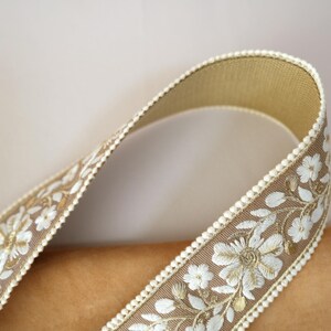 May include: Close-up of a decorative ribbon with a floral design. The ribbon is a light brown color with white embroidered flowers and leaves. The edges are trimmed with a cream-colored beaded border. The ribbon is curved, showing its texture and detail.
