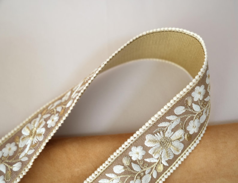 May include: Close-up of a decorative ribbon with a floral design. The ribbon is a light brown color with white embroidered flowers and leaves. The edges are trimmed with a cream-colored beaded border. The ribbon is curved, showing its texture and detail.