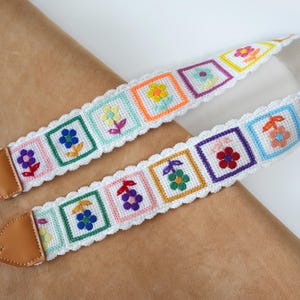 May include: A colorful camera strap with a white base, featuring embroidered floral designs in square frames. The strap has a scalloped edge and brown leather ends. The flowers are in various colors, including yellow, blue, and pink.