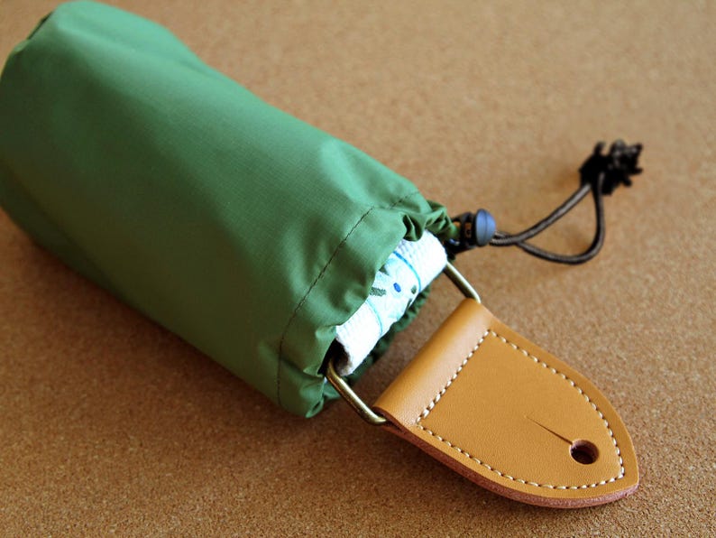 May include: A green drawstring bag with a brown leather strap and a metal buckle. The bag is rolled up and tied with a black cord.