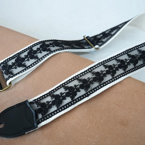 May include: A white guitar strap featuring a black floral lace design. The strap has black leather ends and gold-coloured metal hardware. The lace pattern is a repeating floral motif.