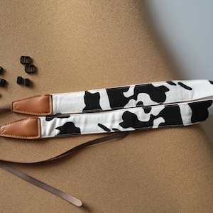 May include: A black and white cow print camera strap with brown leather accents. The strap is adjustable and has a detachable design.