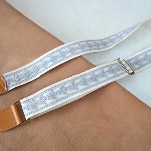 May include: A guitar strap featuring a cream-coloured background with a light blue floral pattern. The strap has brown leather ends and a brass-coloured metal buckle. The strap is designed to be worn over the shoulder.