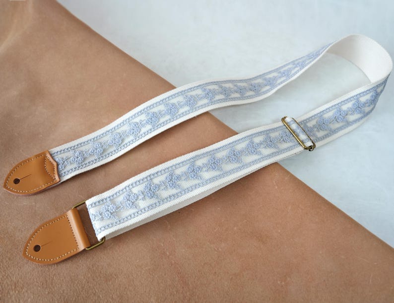 May include: A guitar strap featuring a cream-coloured background with a light blue floral pattern. The strap has brown leather ends and a brass-coloured metal buckle. The strap is designed to be worn over the shoulder.