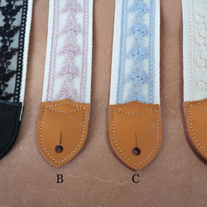 May include: Four guitar straps with decorative lace designs. The straps feature white fabric with lace in black, lavender, light blue, and cream. Each strap has a brown leather end with a hole for attachment. The straps are labelled A, B, C, and D.