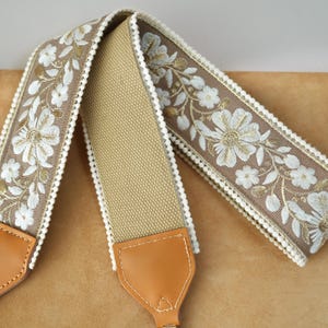 May include: A camera strap with a tan canvas center and brown floral embroidered edges. The floral design features white flowers and green leaves. The strap has tan leather ends and gold-colored metal hardware.