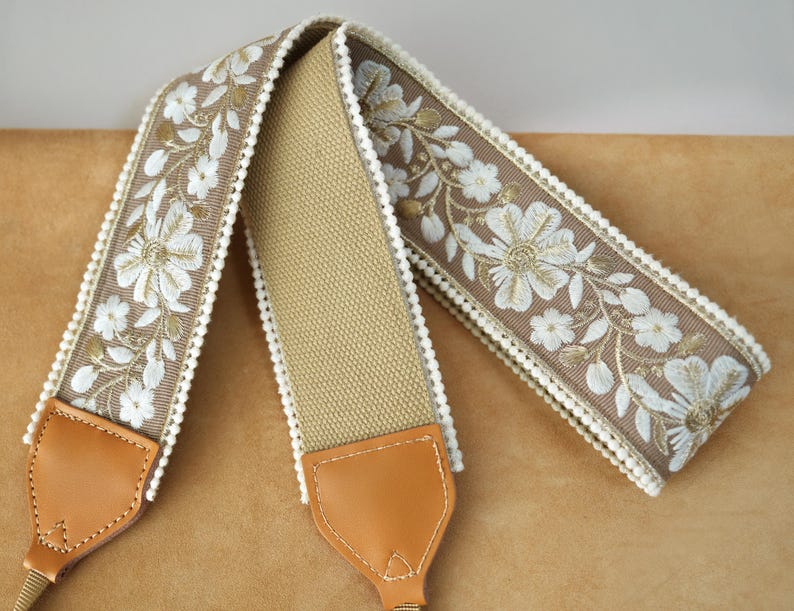 May include: A camera strap with a tan canvas center and brown floral embroidered edges. The floral design features white flowers and green leaves. The strap has tan leather ends and gold-colored metal hardware.