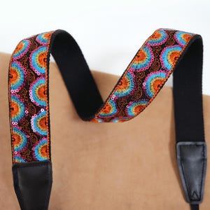 May include: A colorful camera strap with a black backing and leather ends. The strap features a repeating pattern of blue, orange, and pink floral designs on a dark brown background. The strap is designed to be worn over the shoulder.