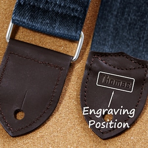 May include: Close-up of a guitar strap with dark denim fabric and brown leather ends. One end features the name "Thomas" in a white rectangle, with "Engraving Position" text below. The strap has silver metal hardware.