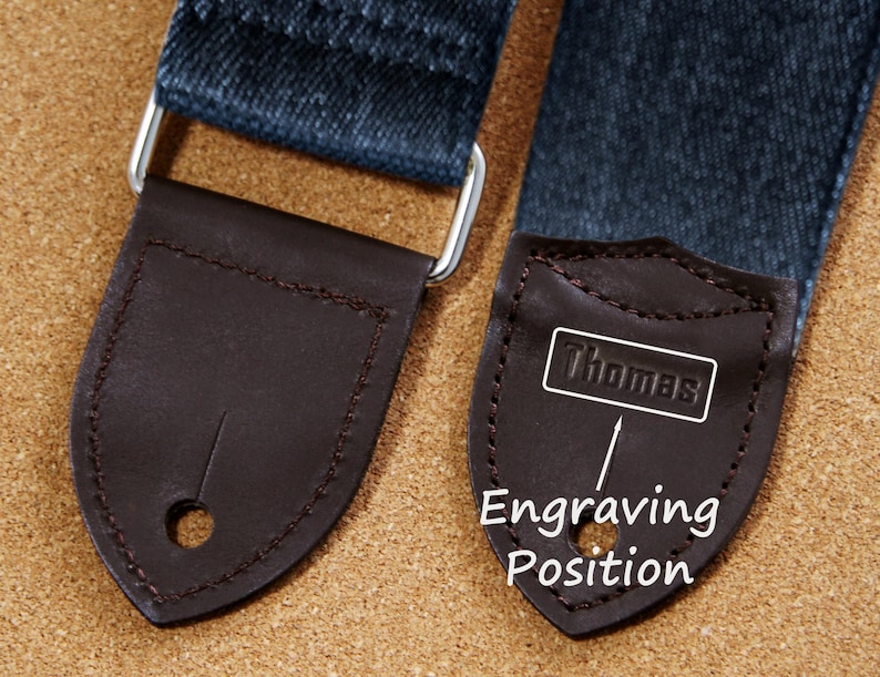 May include: Close-up of a guitar strap with dark denim fabric and brown leather ends. One end features the name "Thomas" in a white rectangle, with "Engraving Position" text below. The strap has silver metal hardware.