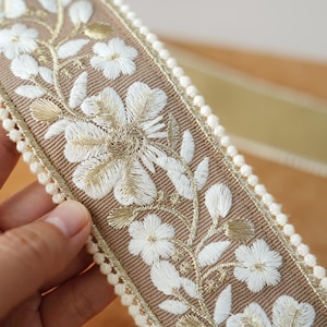 May include: Close-up of a decorative ribbon featuring embroidered white flowers and gold accents on a tan background. The ribbon is trimmed with a cream-colored pom-pom edge, suitable for various craft projects.